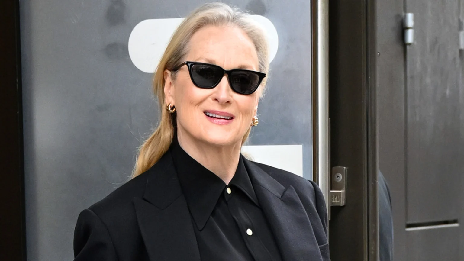 Meryl Streep smiles slightly, wearing black cat-eye sunglasses, gold hoop earrings, a black collared shirt with light buttons, and a black blazer, with her blonde-grey hair pulled back.
