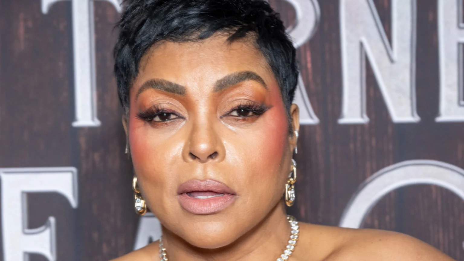 Actress Taraji P. Henson in a close-up portrait, with short dark hair, orange eyeshadow, pink blush, gold hoop earrings, and a sparkly necklace, looking directly at the viewer.