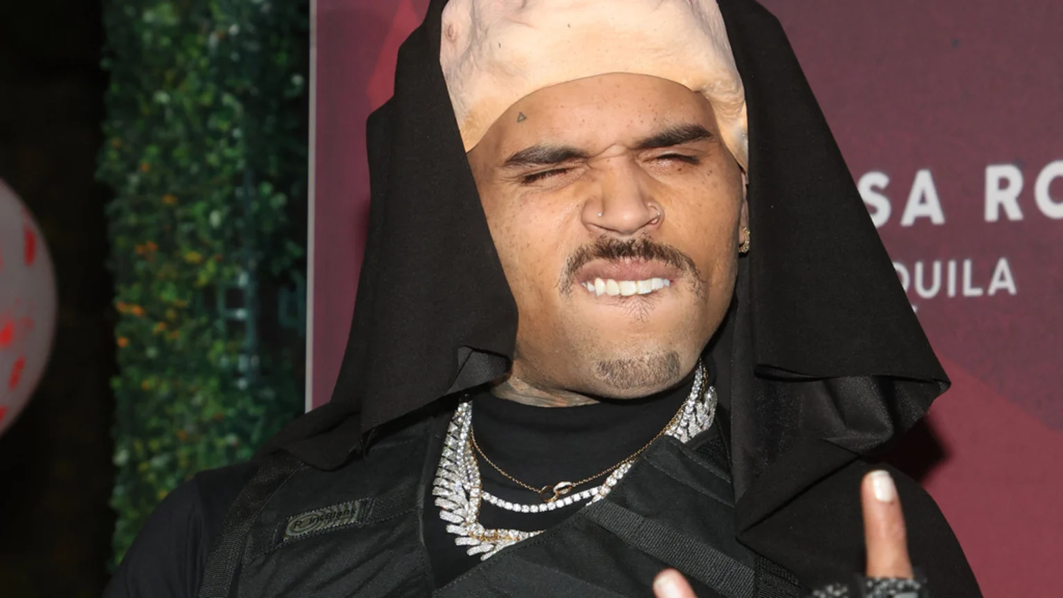 Chris Brown grimaces, wearing a black hood, a light-colored bald cap, a mustache, and multiple diamond necklaces.