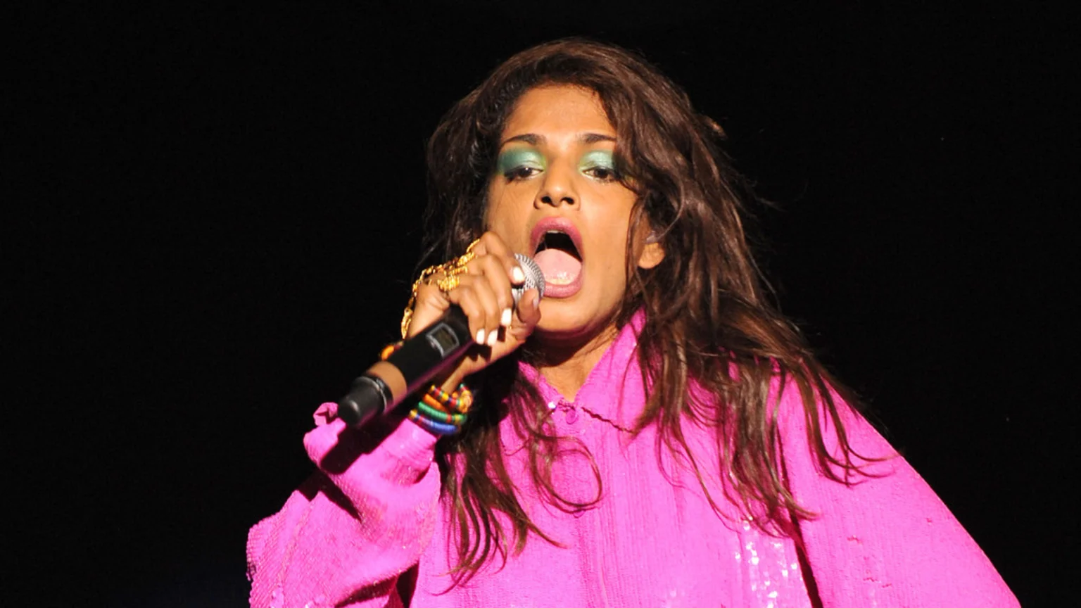 A woman with long brown hair, bright green eyeshadow, and a bright pink sequined top sings into a microphone, her mouth open.