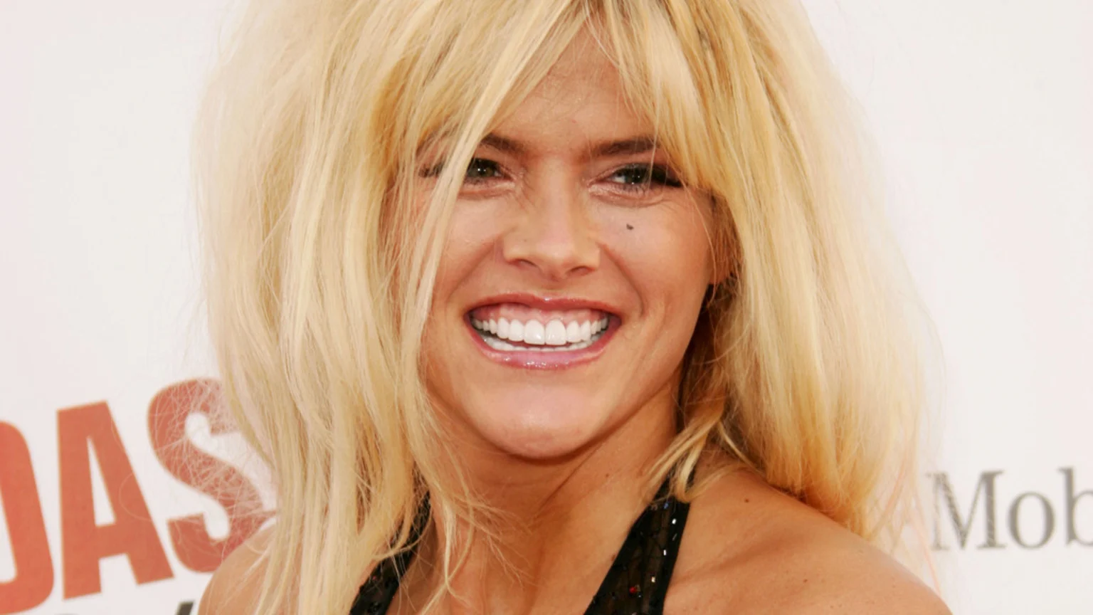 Close-up of a smiling blonde woman with voluminous hair, bangs, a beauty mark near her eye, and a dark top, against a light background with blurred text.