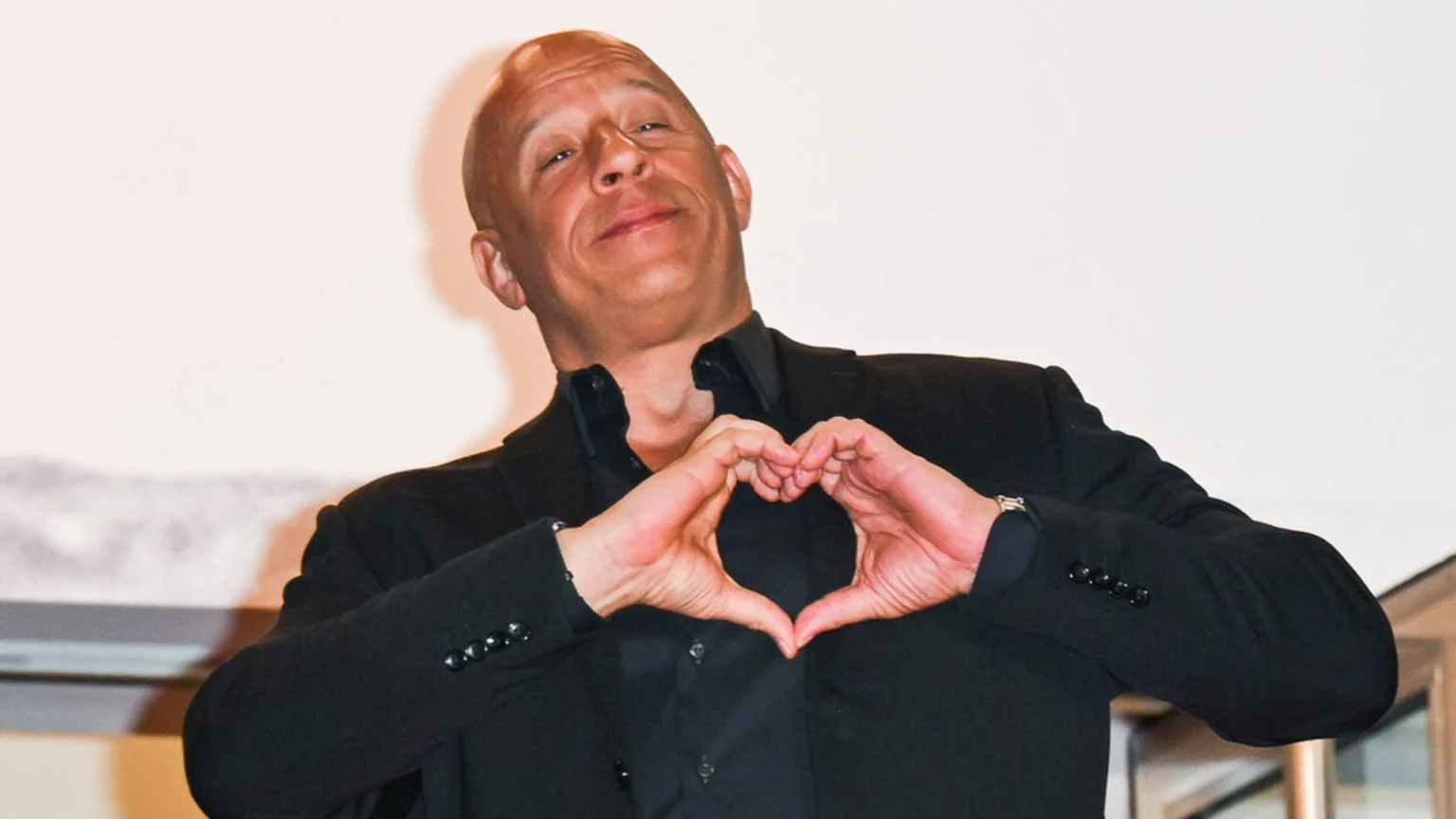 Vin Diesel, wearing a black shirt and blazer, smiles while forming a heart shape with his hands against a light background.