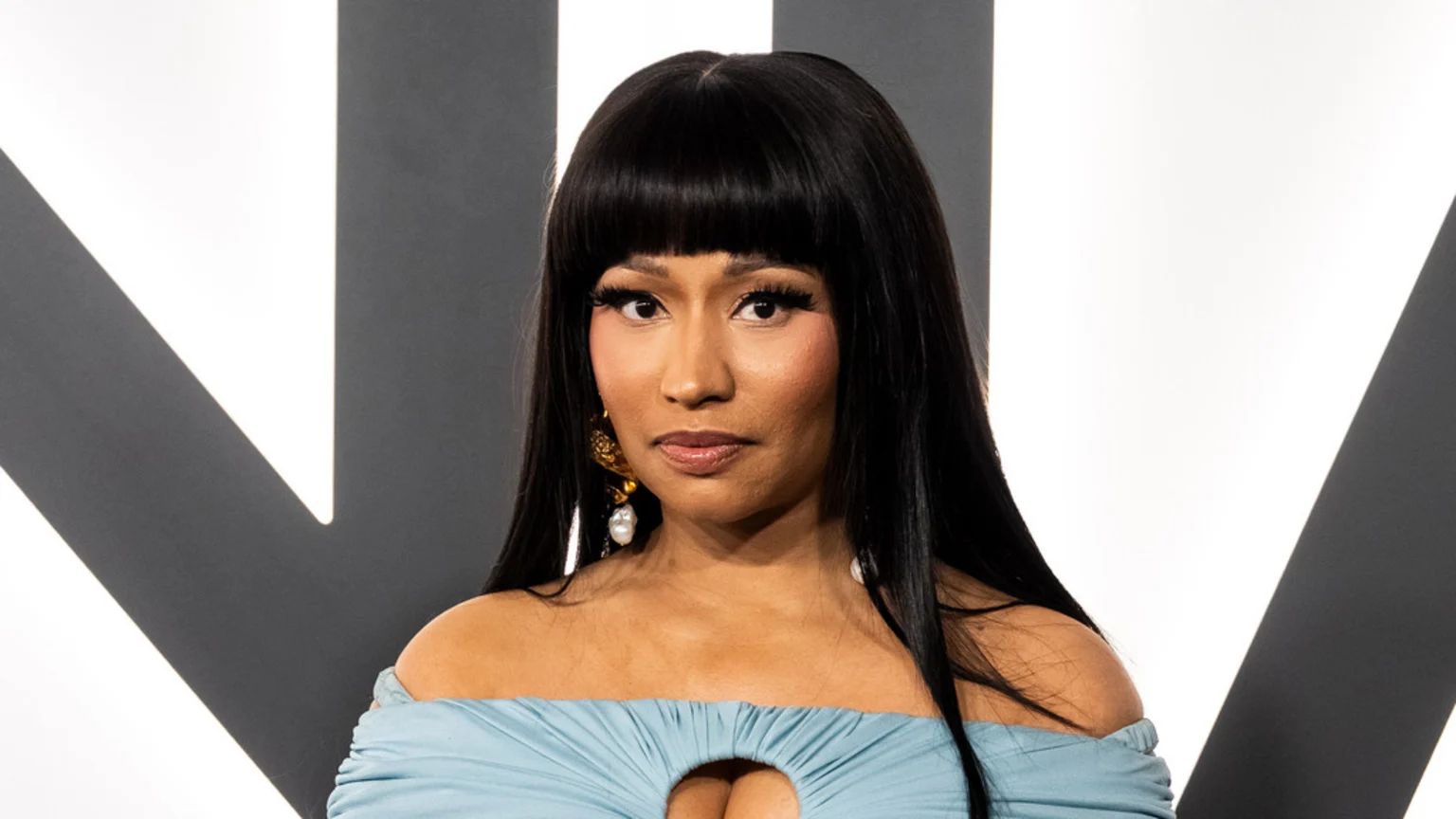 A close-up portrait of Nicki Minaj, with dark hair and bangs, wearing a light blue off-the-shoulder top, ornate earrings, and heavy makeup, against a background of white and dark geometric shapes.