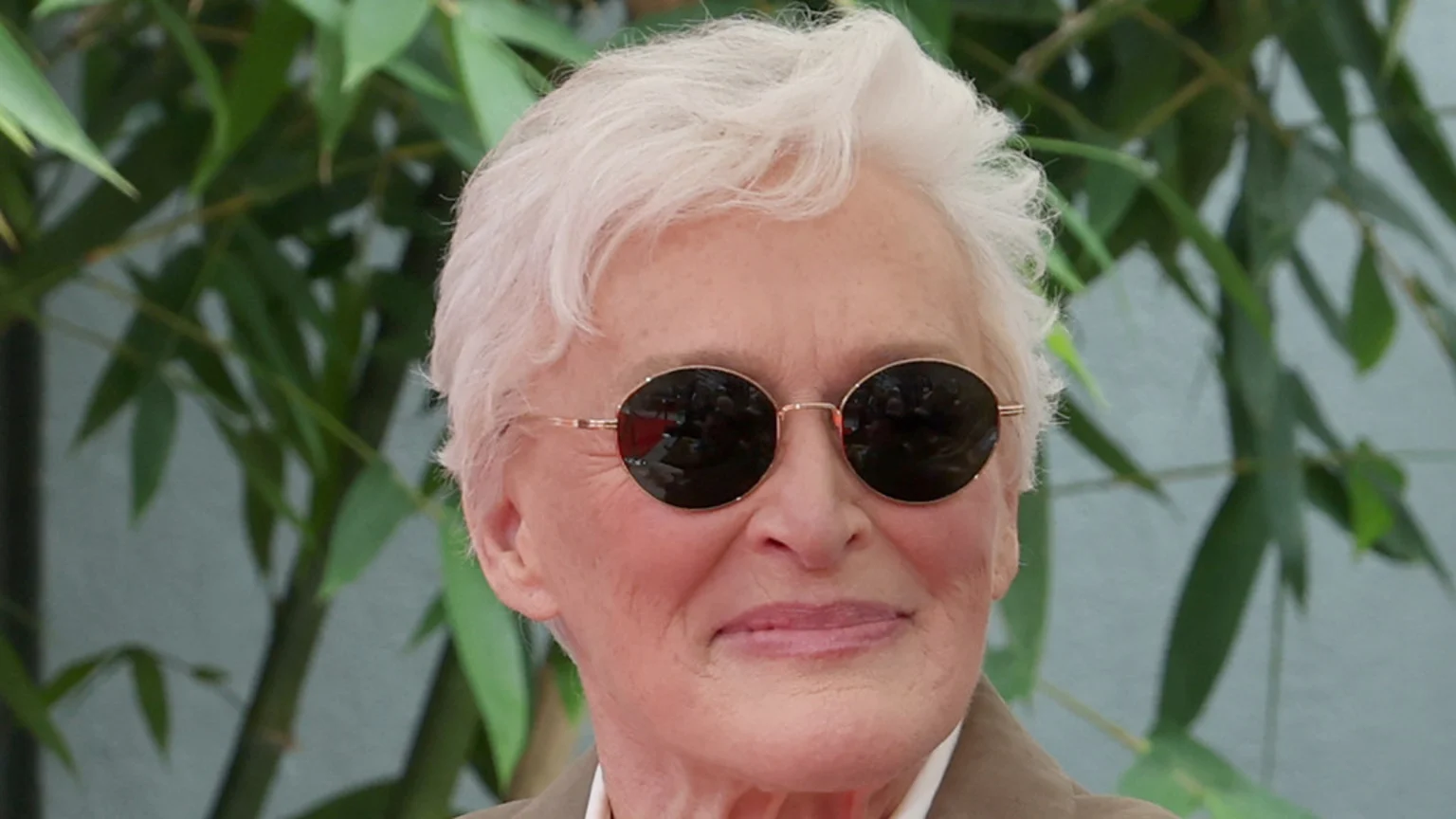 Close-up of Glenn Close with white hair, wearing round dark sunglasses, smiling slightly against a blurred green leafy background.