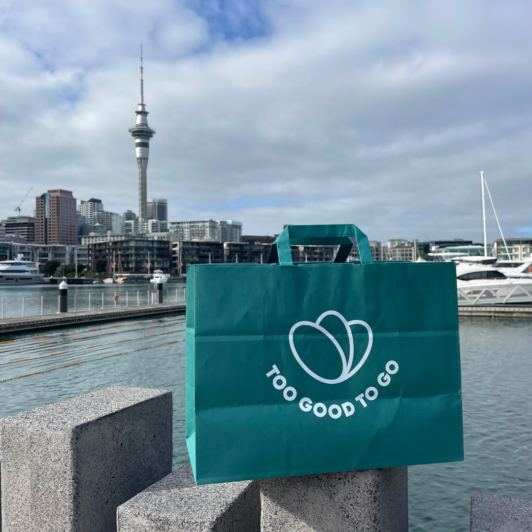 Green 'Too Good To Go' bag with Auckland skyline backdrop. 