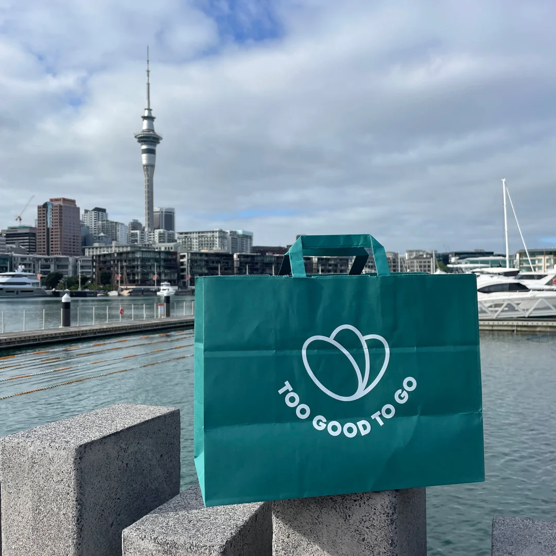 Green 'Too Good To Go' bag with Auckland skyline backdrop.