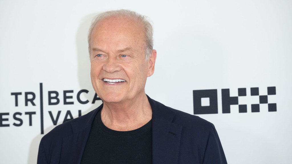 Kelsey Grammer smiles broadly, wearing a dark blue blazer over a black t-shirt, against a white background with "TRIBEC A" text and a geometric logo.