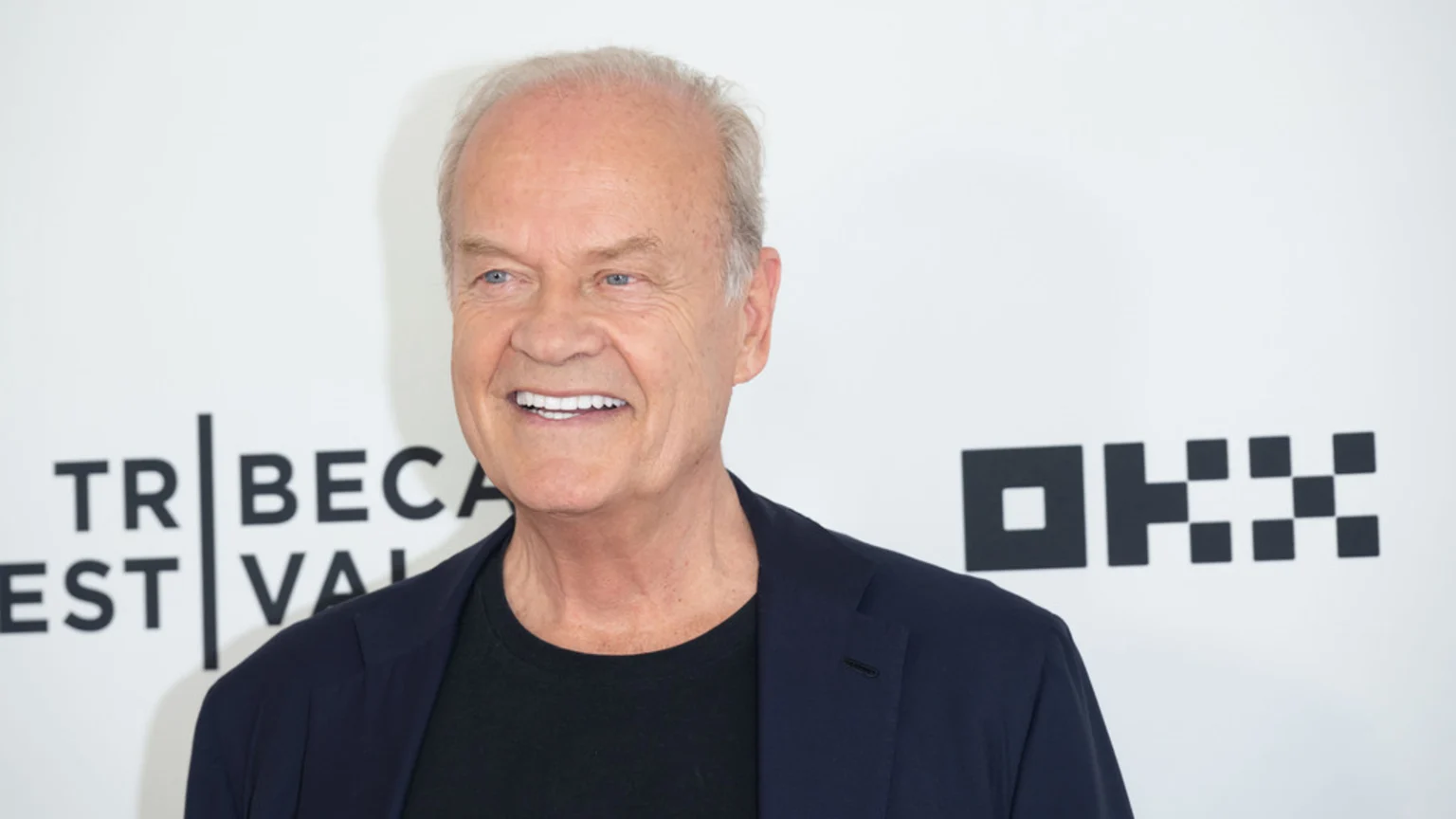 Kelsey Grammer smiles broadly, wearing a dark blue blazer over a black t-shirt, against a white background with "TRIBEC A" text and a geometric logo.