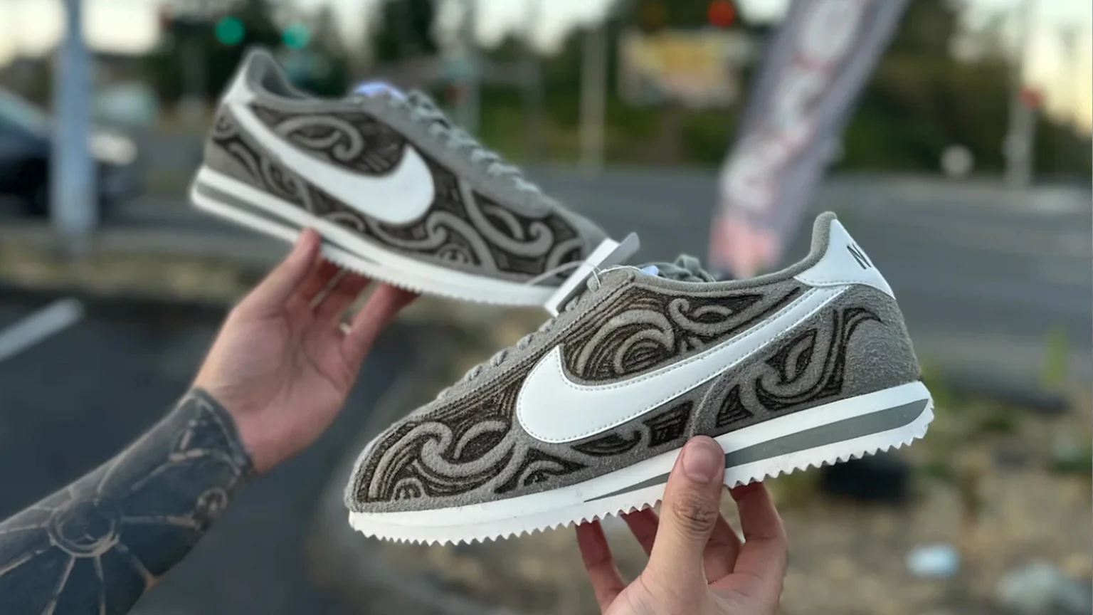 A man's hands holding up a custom pair of Nike cortez sneakers with Māori joru designs burned into the suede textured fabric.