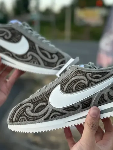 A man's hands holding up a custom pair of Nike cortez sneakers with Māori joru designs burned into the suede textured fabric.