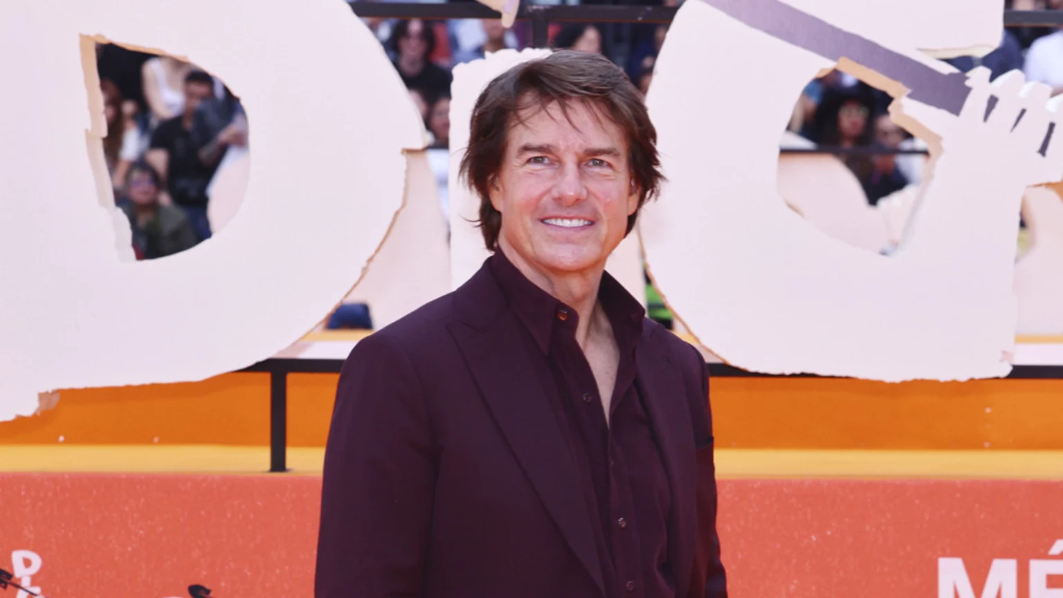 Tom Cruise smiles at the camera in a dark burgundy suit and open-collared shirt, standing before a large white backdrop with stylized letter cutouts.