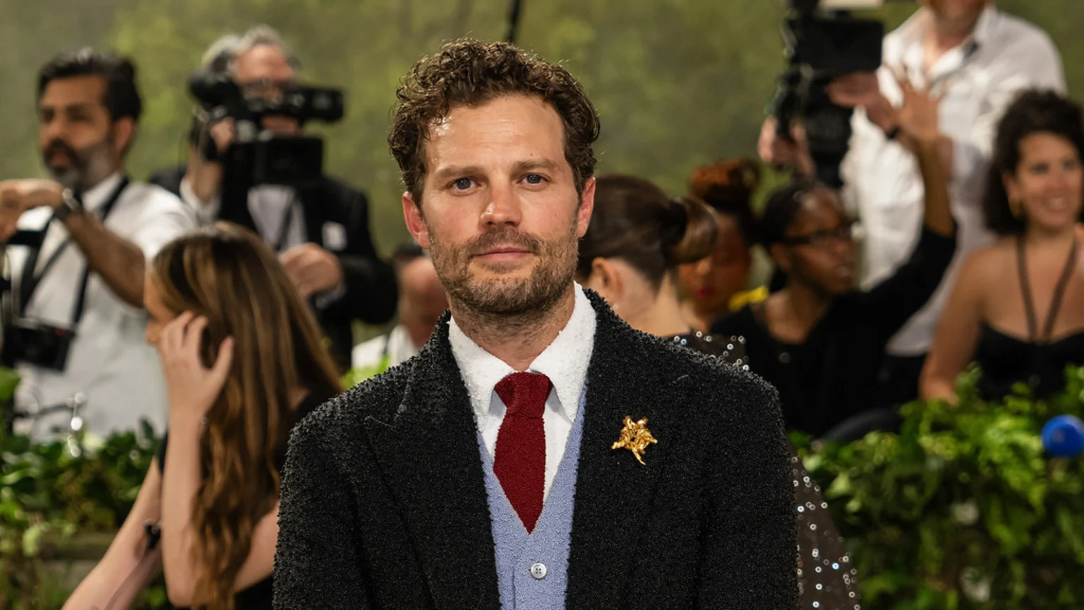 Jamie Dornan looks directly at the camera, wearing a dark textured jacket, a light blue cardigan, a white shirt, a red tie, and a gold brooch, with blurred people and green foliage in the background.