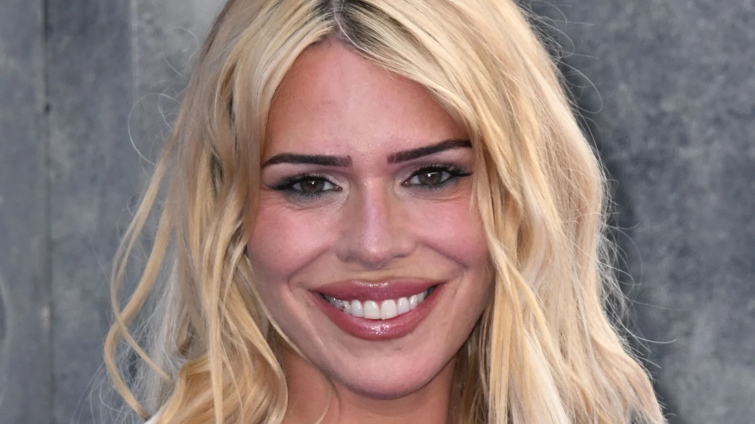 A close-up shot of a blonde woman smiling, featuring her long wavy hair, dark eye makeup, and glossy pink lips, set against a blurry grey background.