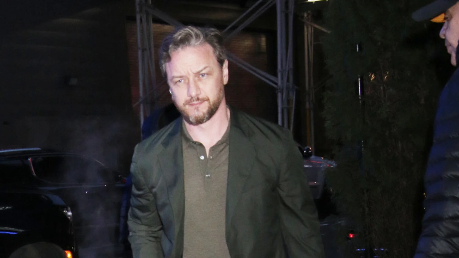 James McAvoy with a beard and grey hair looks directly at the camera, wearing a dark green jacket and polo shirt, against a dark, blurry background.