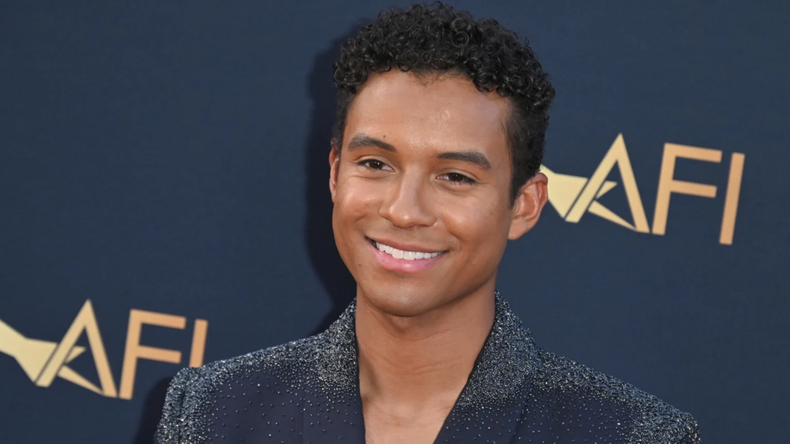 A smiling young man with dark curly hair wears a sparkling dark suit jacket, standing against a dark background with a partial gold AFI logo.