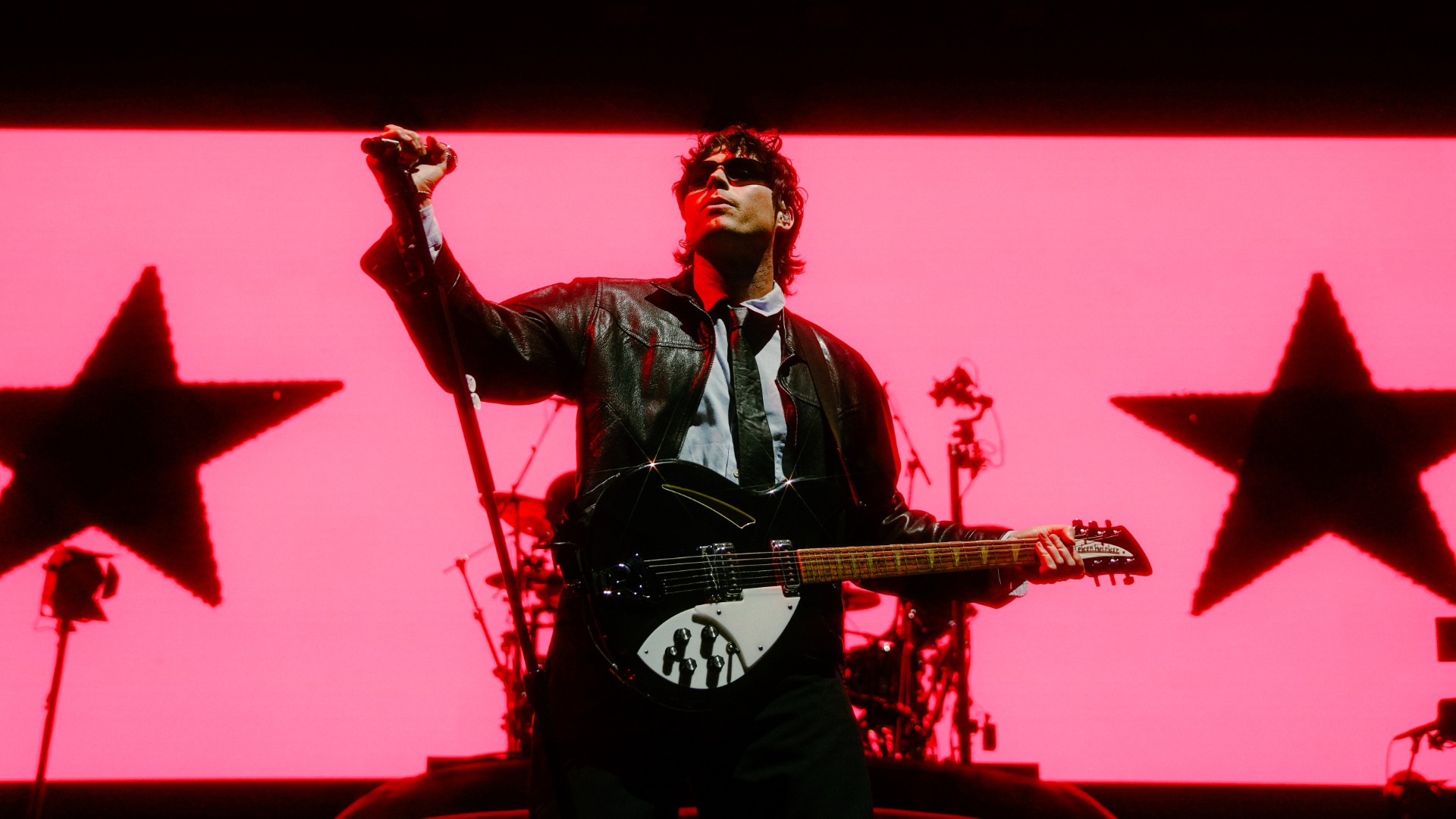 A musician in sunglasses and a leather jacket plays a black guitar on a stage with a bright pink background featuring two large, dark star silhouettes.
