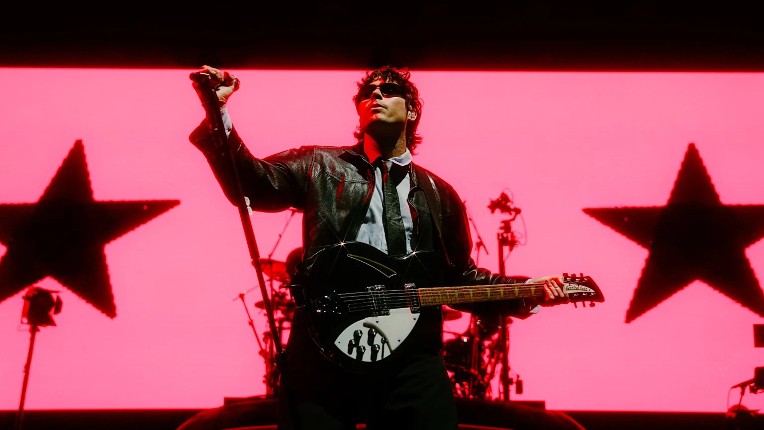 A musician in sunglasses and a leather jacket plays a black guitar on a stage with a bright pink background featuring two large, dark star silhouettes.
