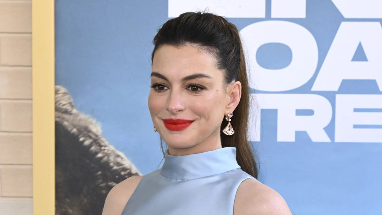 Anne Hathaway smiles softly, wearing a light blue sleeveless top, red lipstick, and earrings, with her hair in a high ponytail against a blurry blue background.