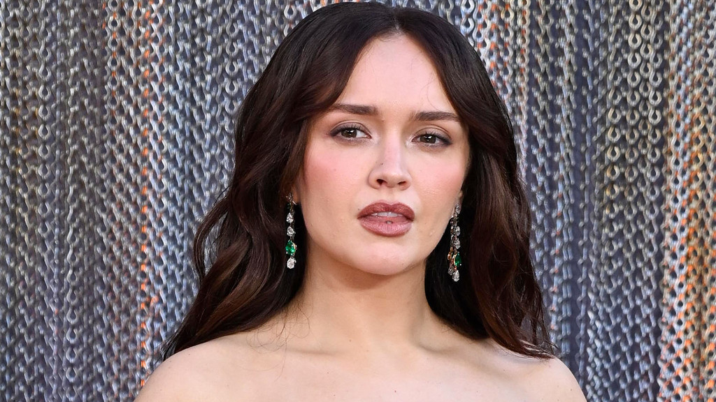 Olivia Cooke, with dark wavy hair and long green and clear earrings, looks at the camera against a textured gray, blue, and orange background.