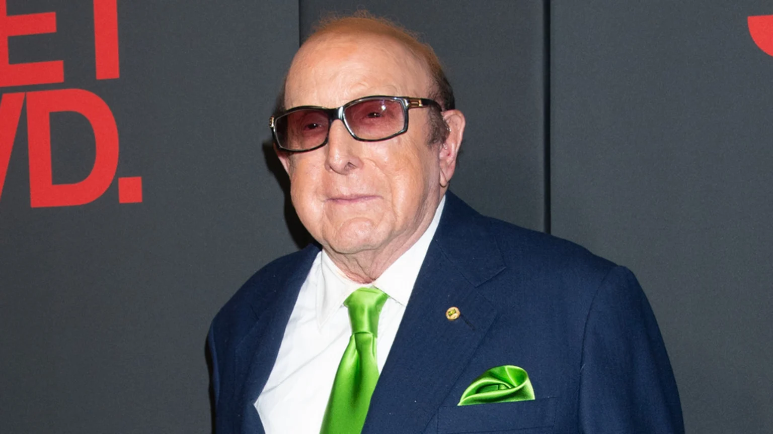 An older man wearing dark sunglasses, a navy suit, a bright green tie, and a matching pocket square stands against a dark background with red text.