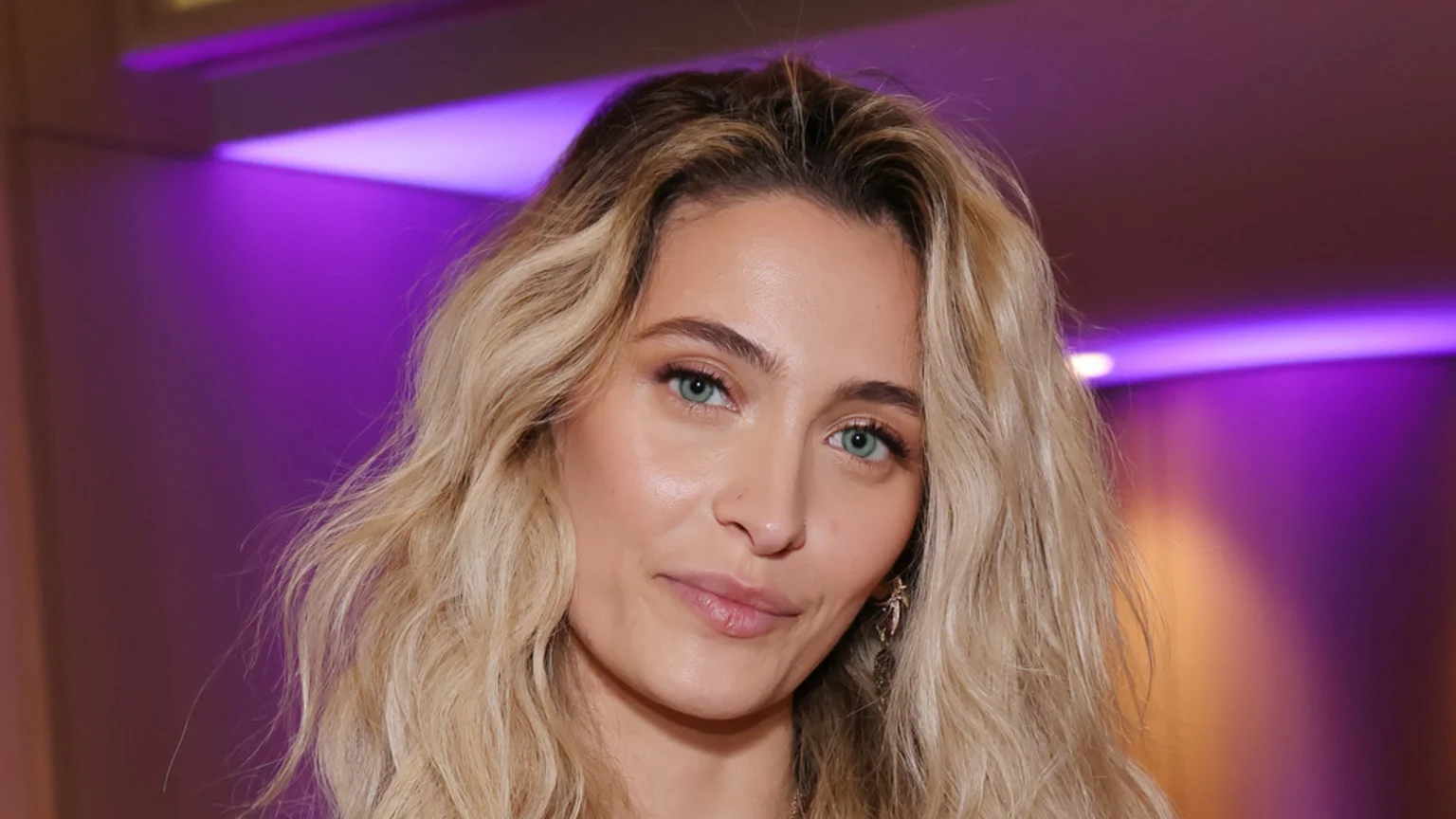 Close-up of Paris Jackson, a woman with long wavy blonde hair, blue eyes, and a nose piercing, looking subtly toward the viewer with a slight smile.