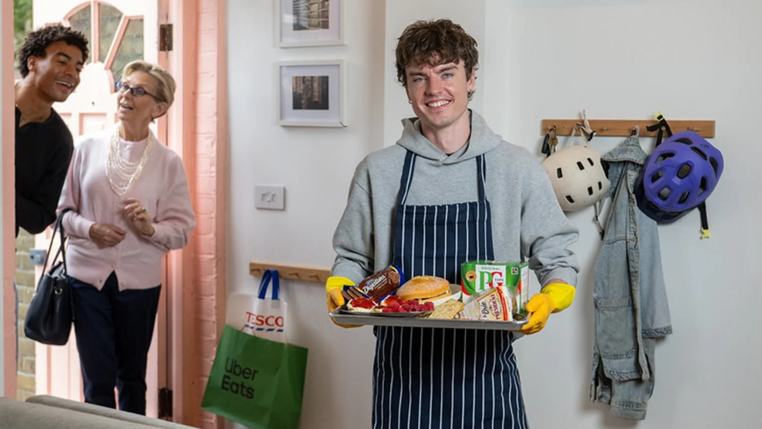 A smiling man in an apron holds a tray of food while two guests arrive at a pink doorway, with grocery bags nearby.