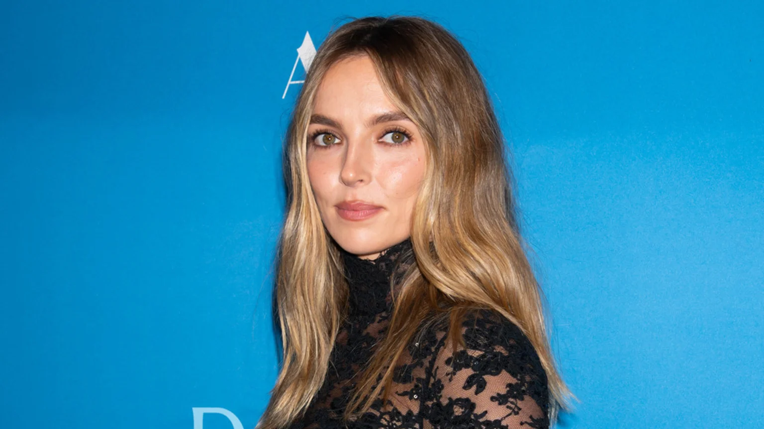 Jodie Comer, with long wavy blonde hair and a sheer black lace top, looks directly at the viewer against a bright blue background.