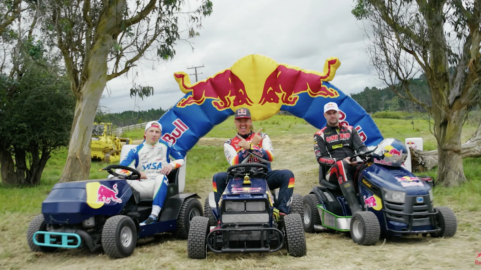 Three racing drivers sit on blue Red Bull-branded lawnmowers under a large inflatable Red Bull arch in a grassy outdoor setting, with the middle driver making a hand gesture.
