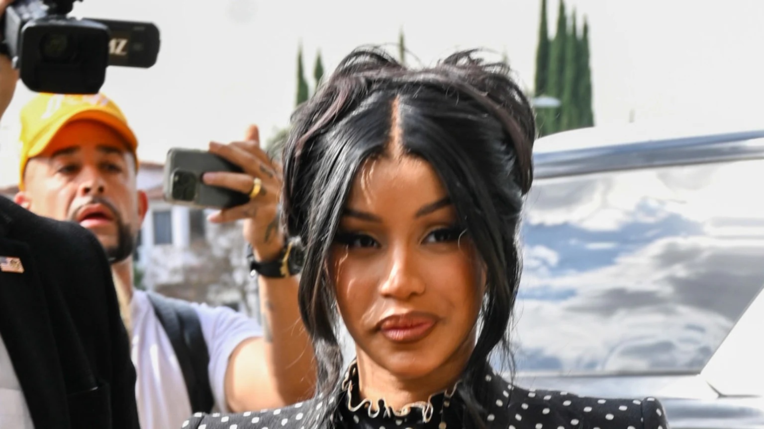 Cardi B, a woman with dark hair in an updo and a polka-dot top, looks forward as men behind her capture photos and video.