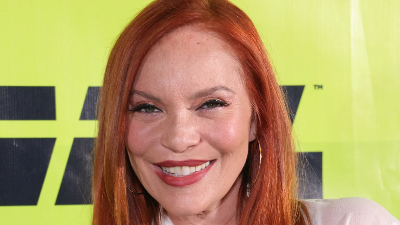 A smiling woman with long reddish-orange hair, prominent eye makeup, and red lipstick, wearing hoop earrings, poses against a neon green background with a dark logo.