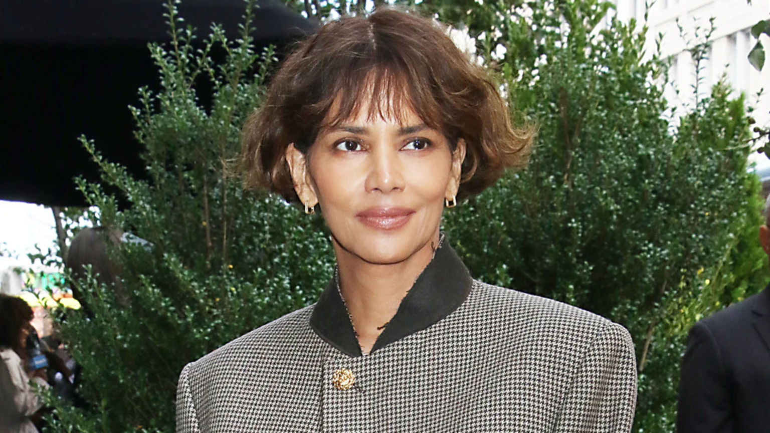 Halle Berry smiles subtly, wearing a houndstooth jacket with a dark collar and a gold brooch, against a background of green foliage.