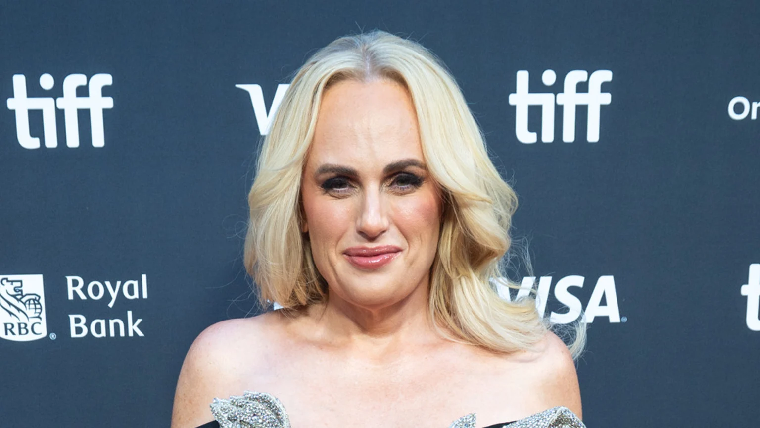 Rebel Wilson, a blonde woman, smiles softly, wearing a strapless sparkly top against a dark event backdrop with "tiff" and "RBC Royal Bank" logos.