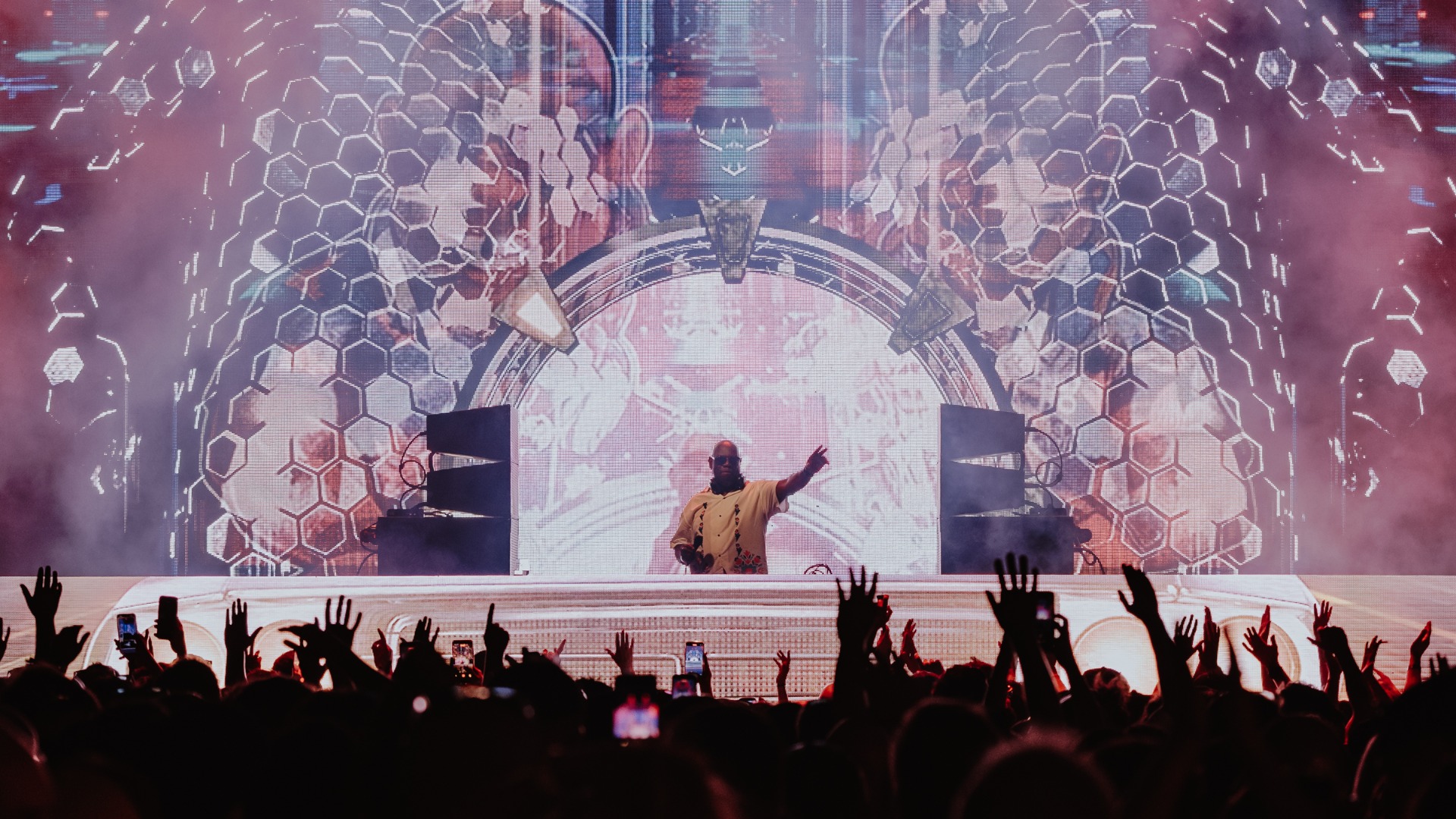 A DJ with a bald head and light-colored shirt performs on a smoky stage in front of a large LED screen displaying vibrant hexagonal patterns, with a silhouetted crowd raising their hands in the foreground.
