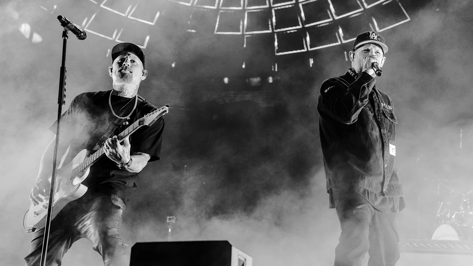 Black and white photo of two men performing on a smoky stage with a patterned light backdrop; one plays electric guitar, the other sings into a microphone.