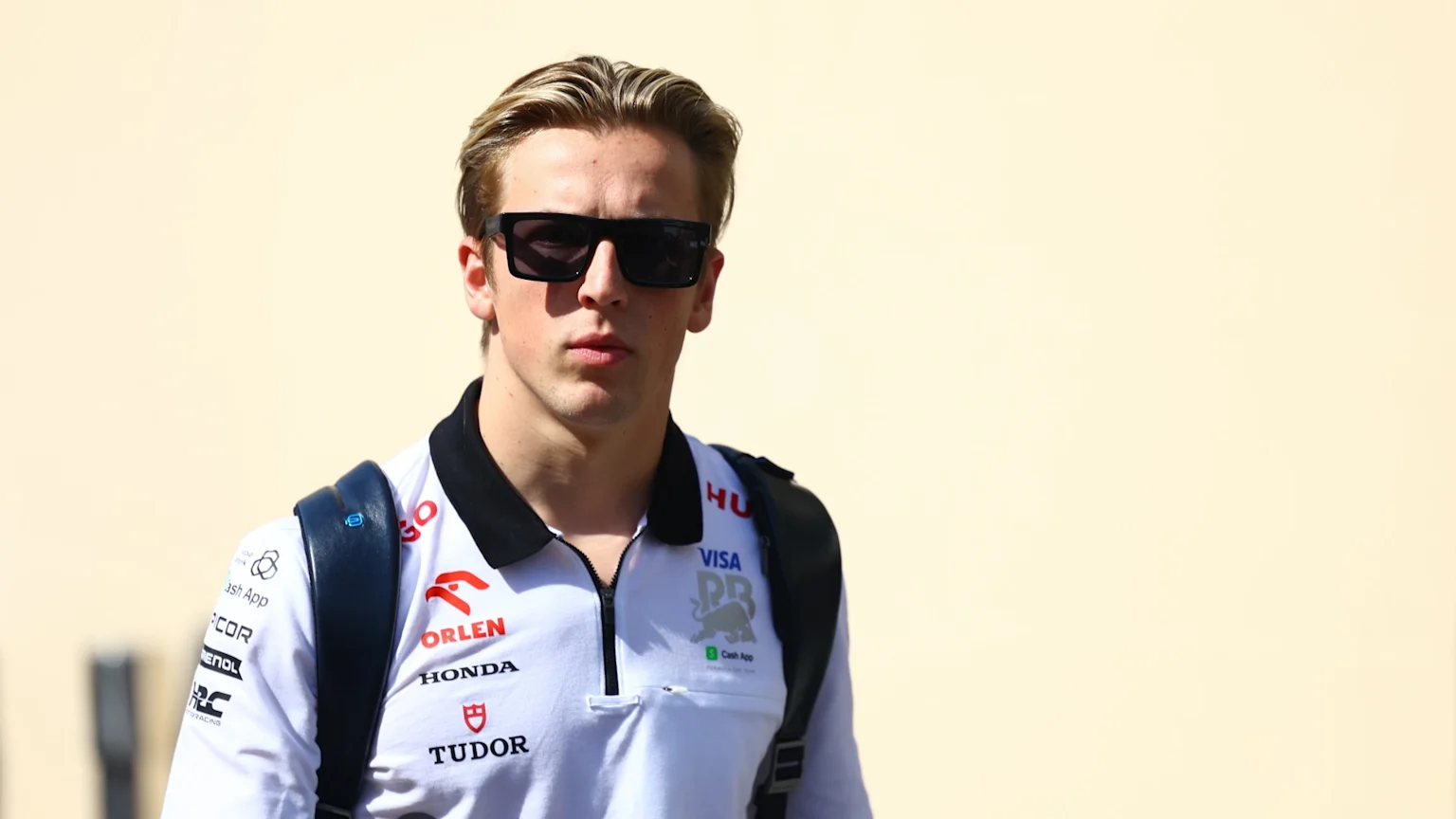 A young man with blonde hair and sunglasses wears a white polo shirt with motorsport sponsor logos, looking forward outdoors.