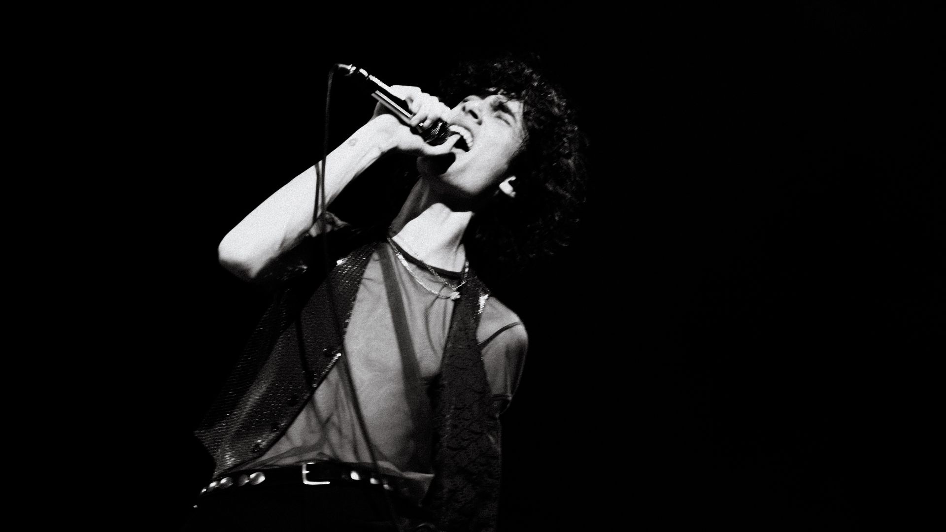 A black and white, low-angle shot of a male singer with curly hair, wearing a sheer top and sequined vest, passionately singing into a microphone, head tilted back.