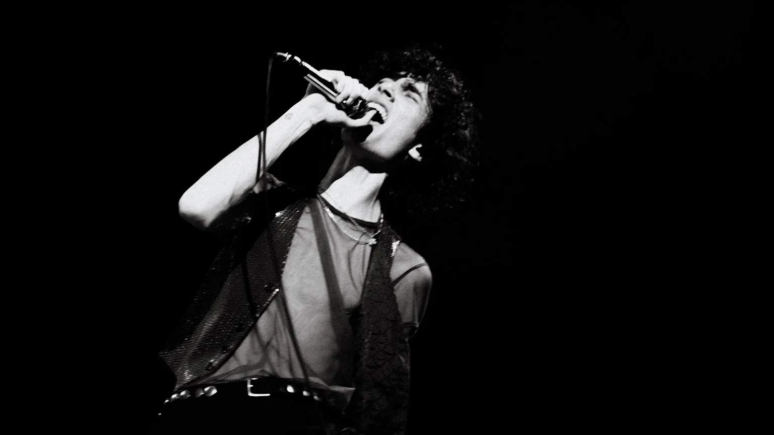 A black and white, low-angle shot of a male singer with curly hair, wearing a sheer top and sequined vest, passionately singing into a microphone, head tilted back.