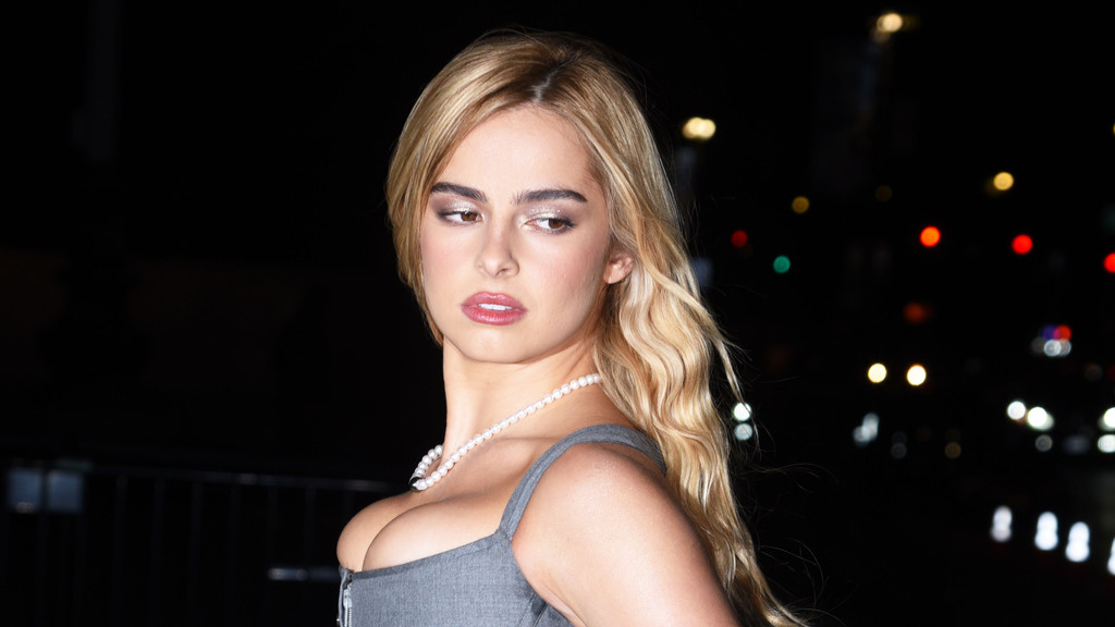 A blonde woman with shimmery eye makeup, wearing a pearl necklace and a grey top, looks left against a dark, blurred background of city lights.