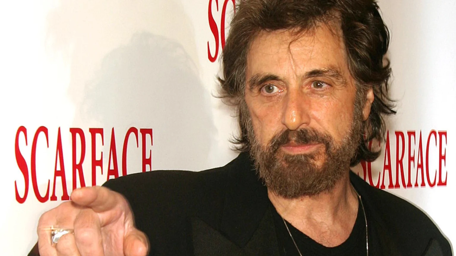 A bearded Al Pacino points his hand forward, standing against a white background with repeated red "SCARFACE" logos.