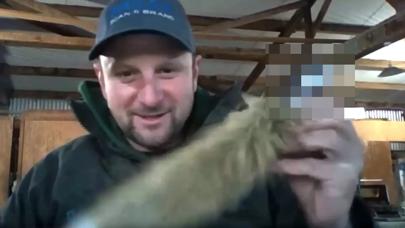 A smiling man in a baseball cap and jacket holds a blurry, furry, light brown object directly in front of him, with a pixelated background.