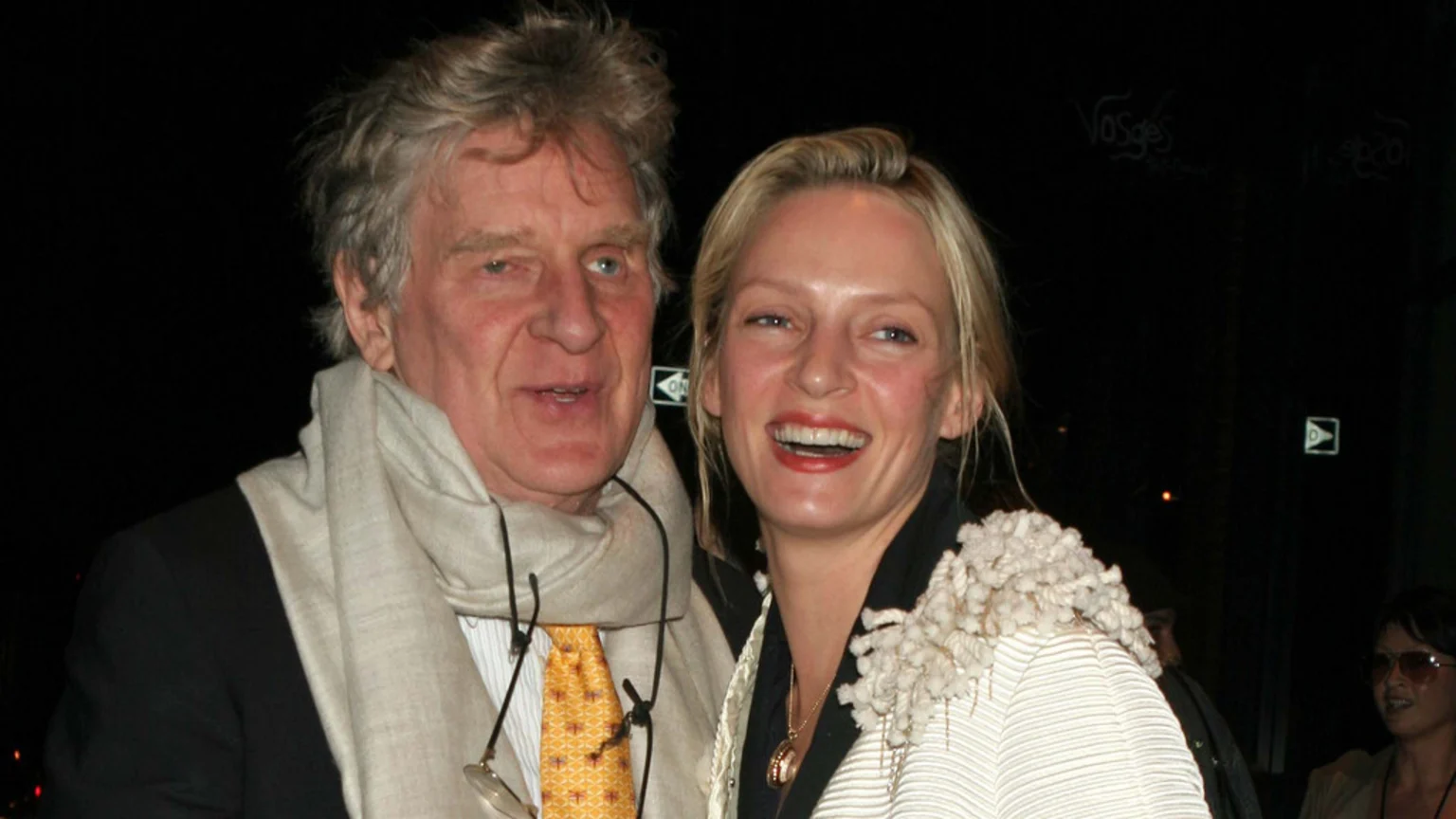 Robert Thurman and Uma Thurman smile broadly, with Robert wearing a suit, yellow tie, and light scarf, and Uma in a white textured jacket.