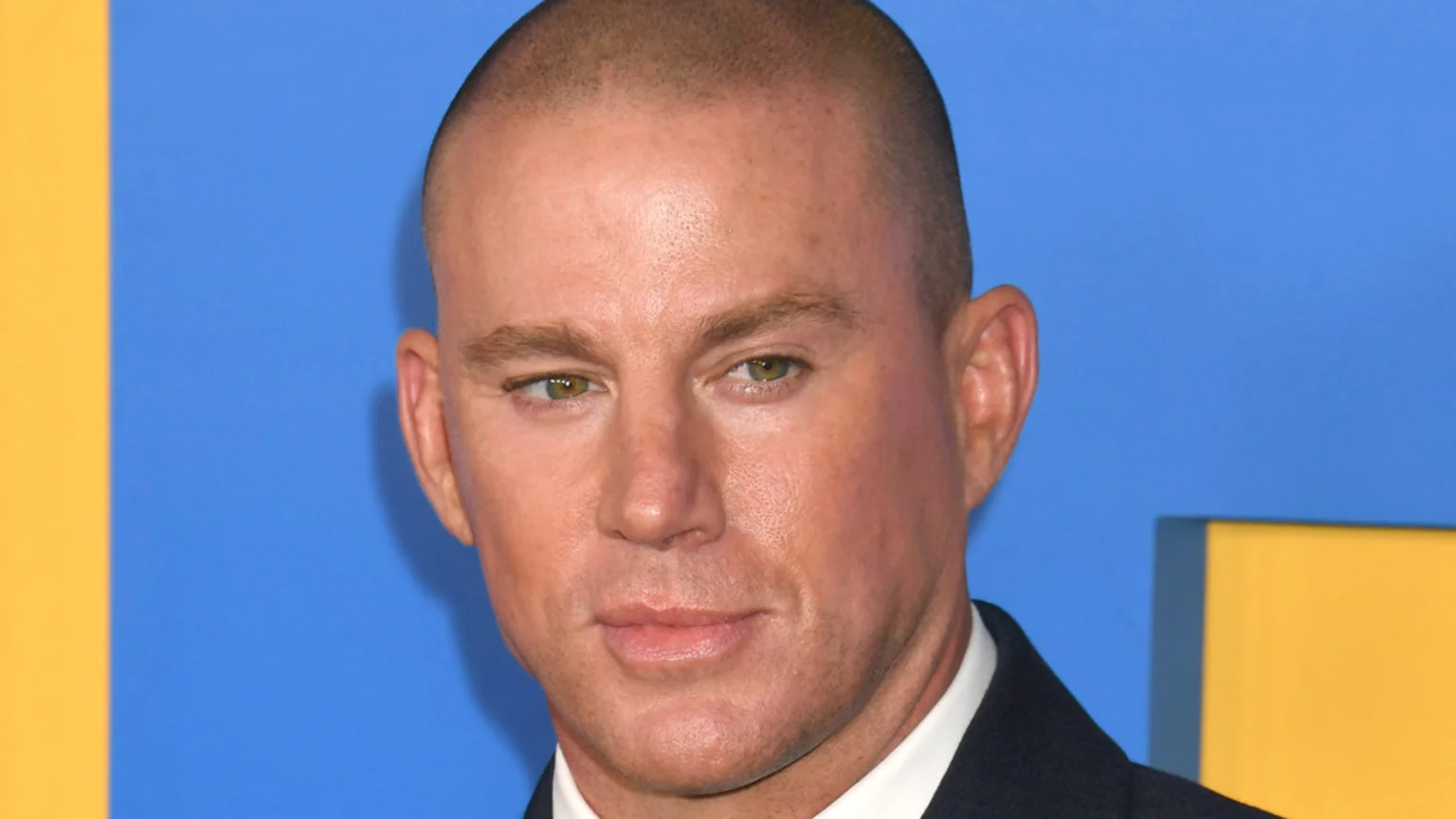 Close-up of actor Channing Tatum with a buzz cut, green eyes, and a slight smile, wearing a dark suit against a blue and yellow background.