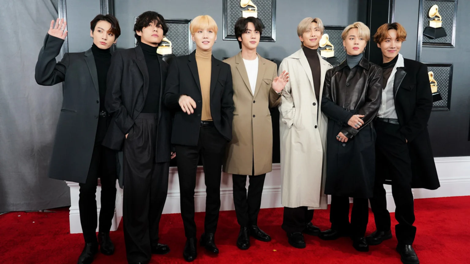 Seven members of BTS stand on a red carpet, posing in long coats and turtlenecks, with golden Grammy award statues visible in the background.