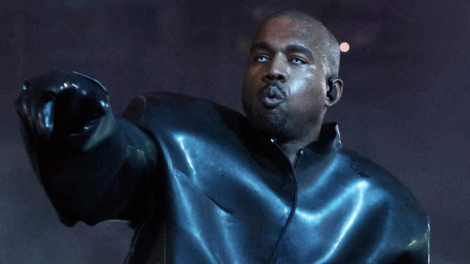 Kanye West, wearing a shiny black jacket and glove with white contact lenses, pursing his lips and raising his left hand on a dark stage.