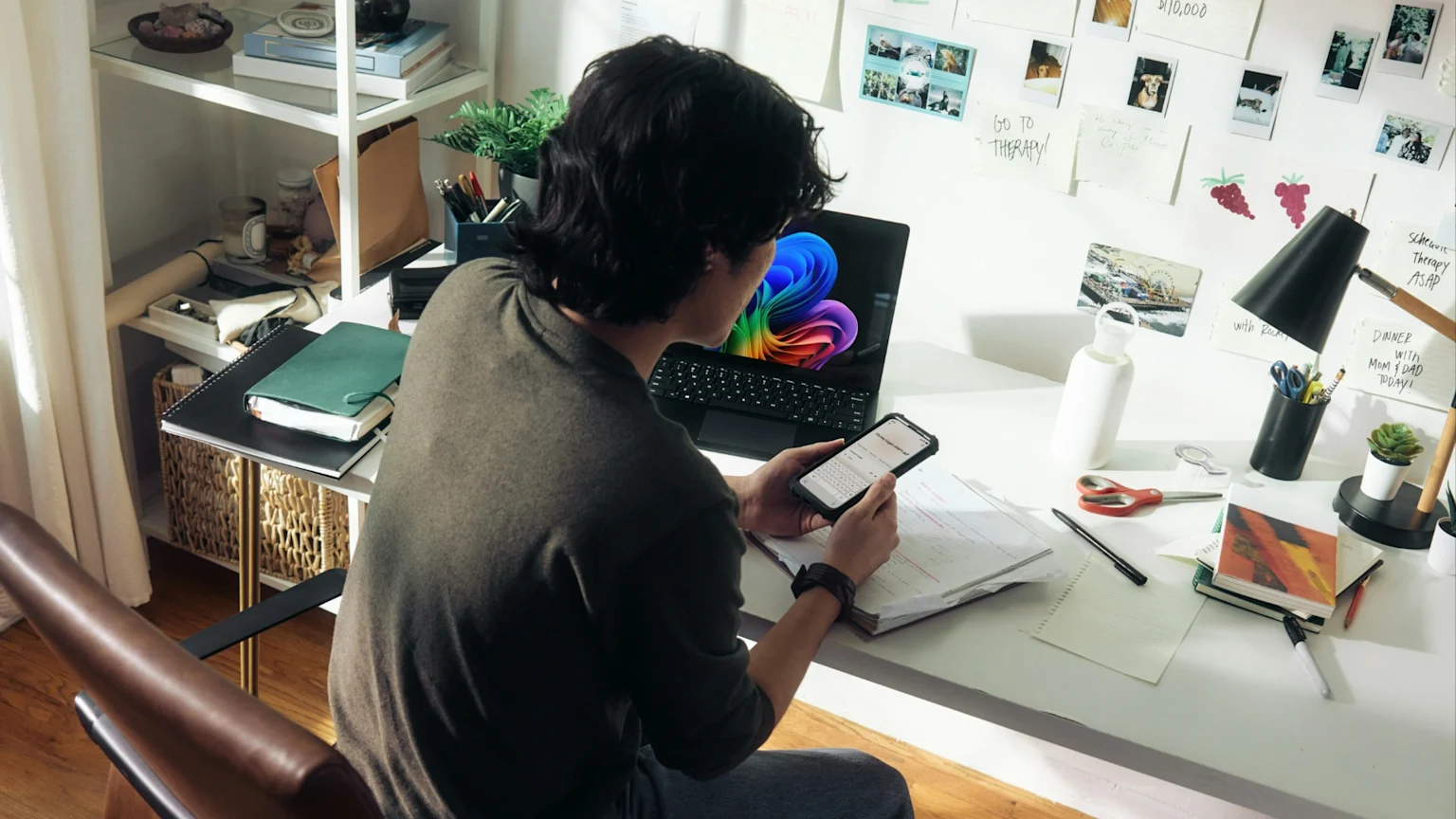 A person with dark hair sits at a brightly lit desk, focused on their smartphone, with a laptop displaying a colorful image, notebooks, and office supplies scattered around.