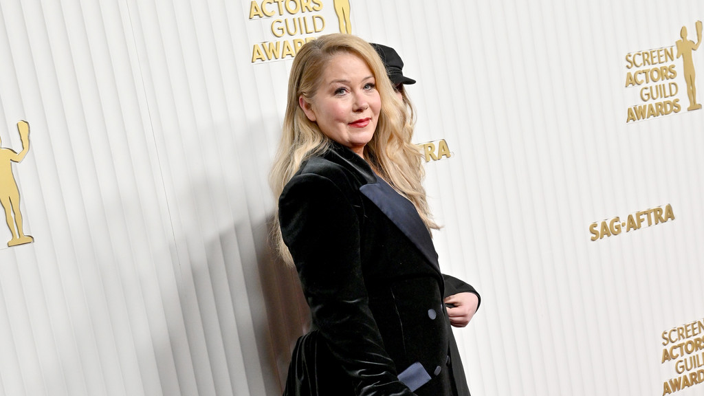 Christina Applegate with long blonde hair, red lipstick, and a dark velvet jacket with a blue lapel poses in front of a white wall with gold SAG-AFTRA logos.