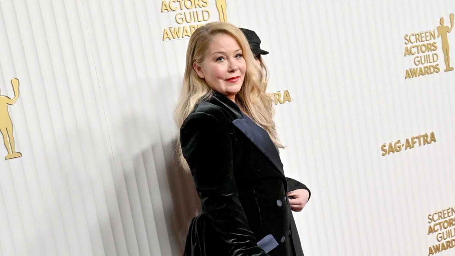 Christina Applegate with long blonde hair, red lipstick, and a dark velvet jacket with a blue lapel poses in front of a white wall with gold SAG-AFTRA logos.
