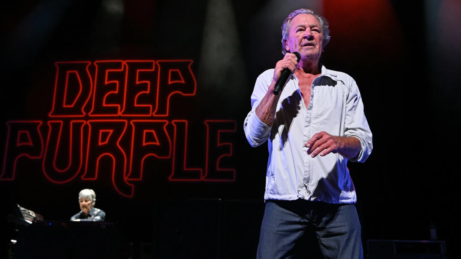 Deep Purple's lead singer stands on stage holding a microphone, with the band's red neon logo prominently displayed behind him and a keyboard player visible to the left.