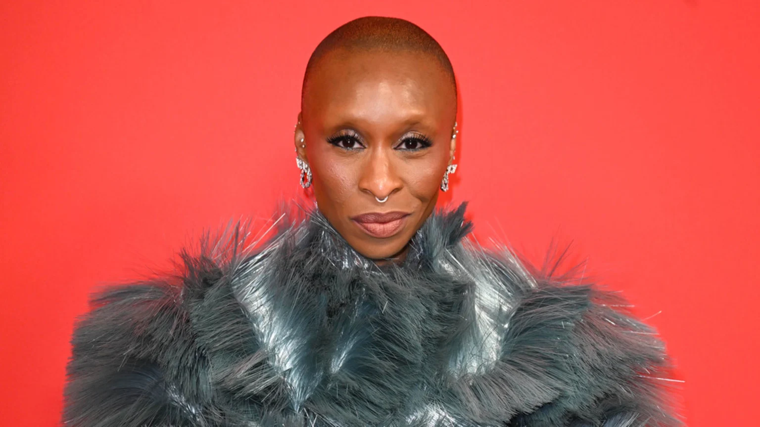 Cynthia Erivo, an actress with a shaved head, wears a grey textured top and a septum piercing, looking directly at the camera against a red background.
