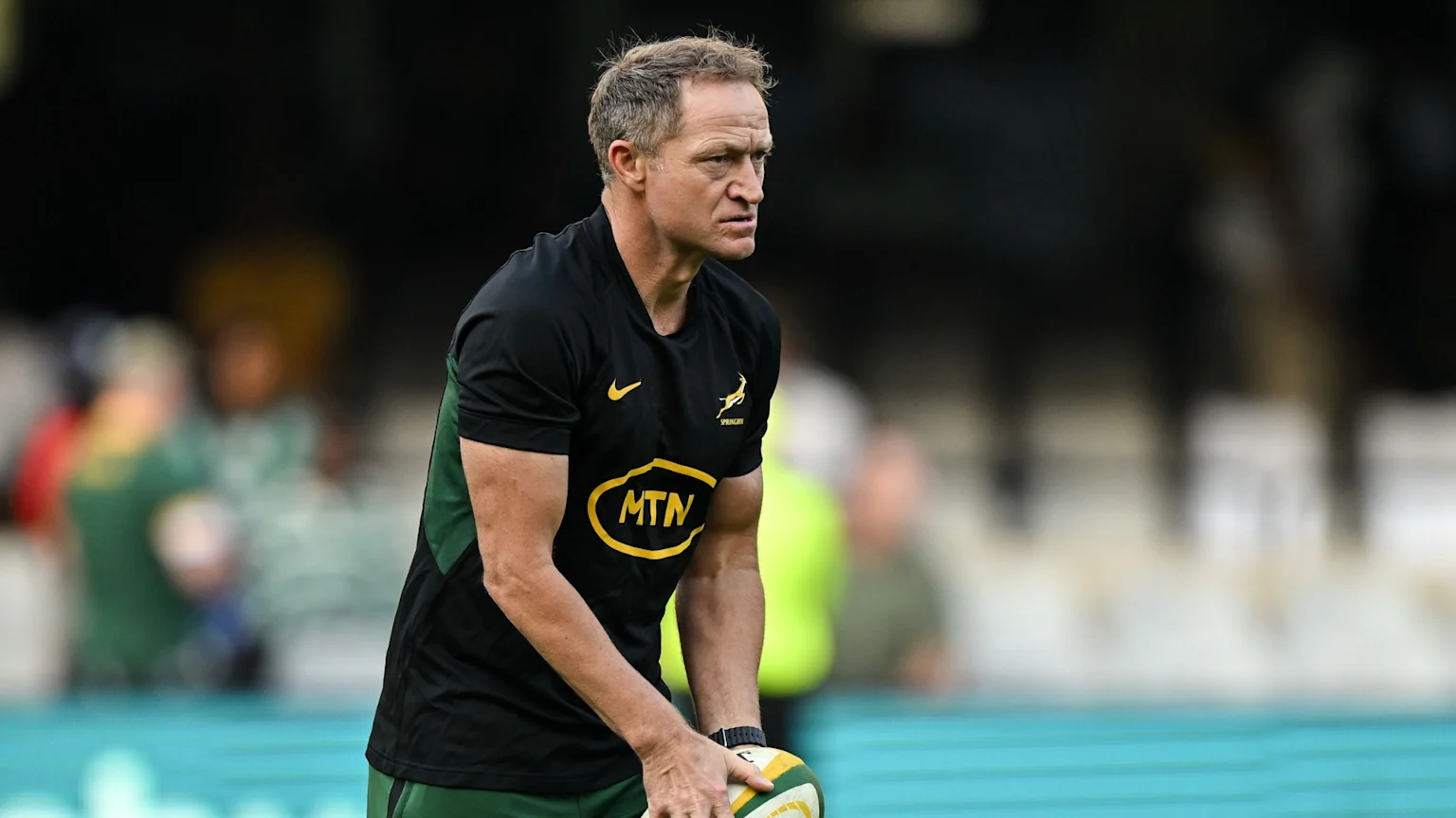 A man wearing a black and green Springboks rugby shirt with "MTN" sponsor logo holds a rugby ball with a focused expression on a blurred field.